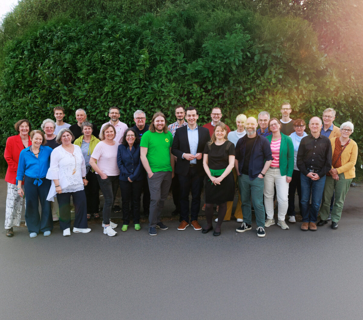 Auf dem Bild stehen viele Menschen unterschiedlichen Alters vor einer grünen Hecke und lächeln in die Kamera.