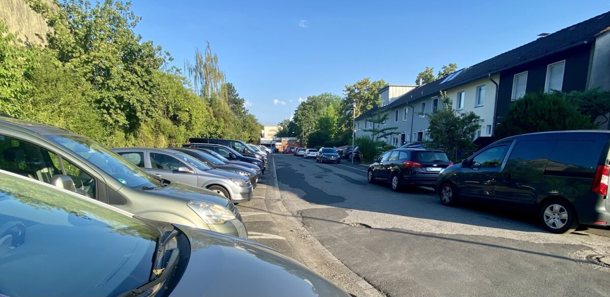 Wohnstraße mit dicht geparkten Autos, Häusern auf der rechten und viel Grün auf der linken Seite.
